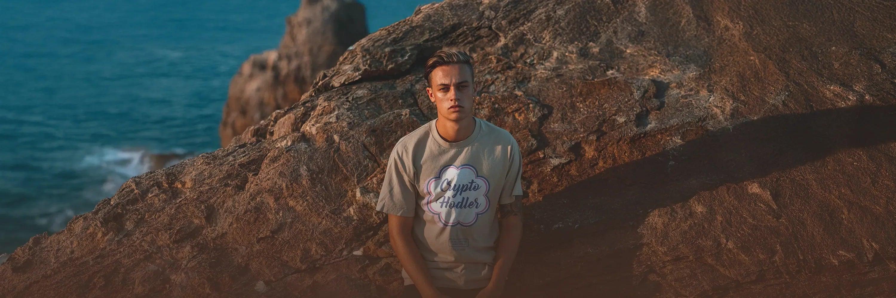 Young man wearing beige oversized "Crypto Hodler" T-Shirt from CUKIDRIP, sitting on coastal rocks – Premium Crypto Streetwear Collection Banner